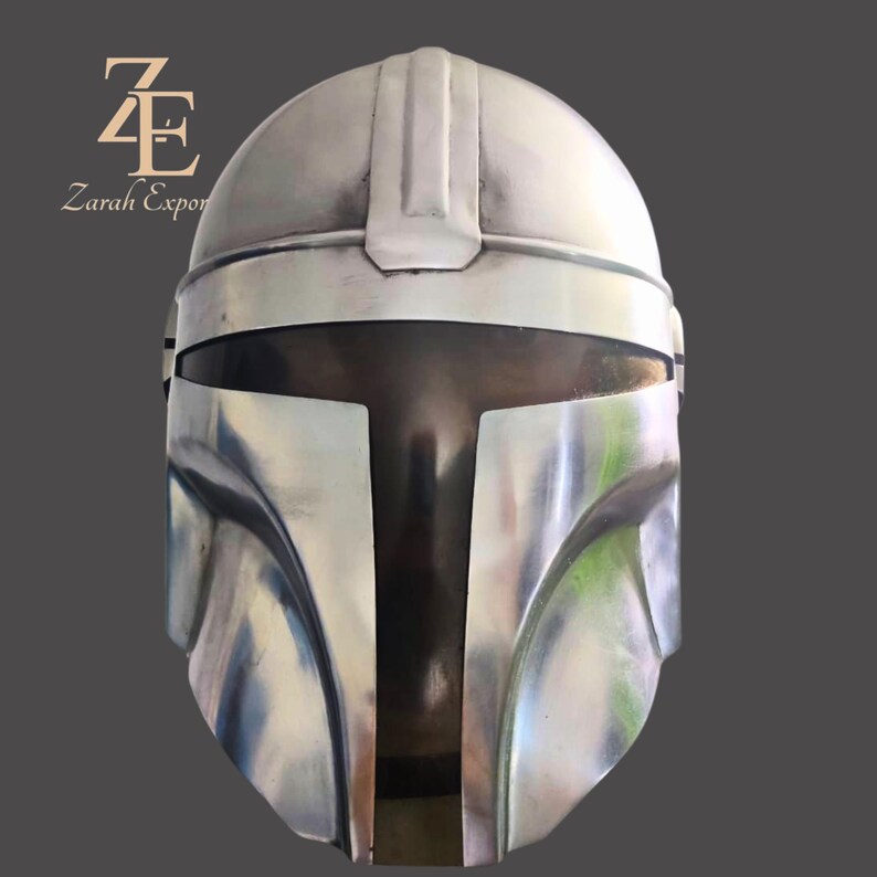 Puede incluir: Un casco mandaloriano plateado con una visera en forma de T y una parte superior redondeada. El casco tiene un acabado met&aacute;lico liso y est&aacute; dise&ntilde;ado para cubrir toda la cabeza. La imagen incluye el texto "Zarah Export" en la esquina superior izquierda.