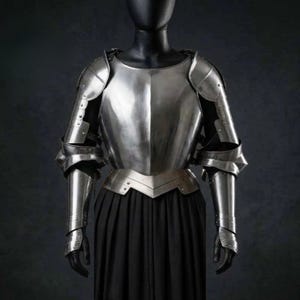 May include: A polished steel breastplate and pauldrons set featuring a central ridge on the chest, articulated shoulder guards, and matching arm defenses. The metallic armor pieces are displayed on a mannequin torso over a long black garment.