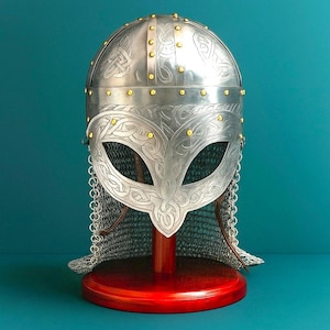 Puede incluir: Un casco vikingo plateado con intrincados grabados de nudos celtas y remaches dorados. El casco presenta una protección facial con aberturas para los ojos y cota de malla. Se exhibe sobre un soporte de madera rojo.
