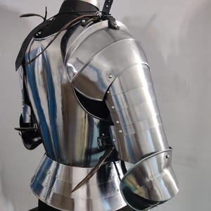 May include: A polished steel suit of armor torso piece featuring a breastplate, a segmented shoulder pauldron, and a full arm guard with a rounded elbow cop. The armor is secured with black leather straps and metal buckles over a dark mannequin.
