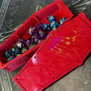 Puede incluir: Una caja de dados roja en forma de ataúd, abierta para revelar una colección de dados poliédricos coloridos. La caja está hecha de un material sólido y tiene una tapa con un acabado reflectante e iridiscente. El fondo presenta una cubierta de libro decorativa.