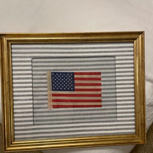 Puede incluir: Bandera americana enmarcada con rayas rojas y blancas y un campo azul de estrellas. La bandera está colocada sobre un fondo a rayas grises y blancas dentro de un marco dorado. Un artículo de decoración patriótica para el hogar.