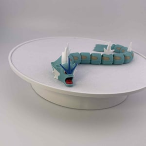 May include: A detailed figurine of a serpentine creature, featuring a teal body with segmented sections and white accents. The creature has a teal head with a red mouth and white spikes. The figurine is displayed on a white, circular platform.