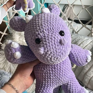 Könnte beinhalten: Ein gehäkeltes Drachen-Stofftier in Lavendel mit weicher, strukturierter Garnoberfläche, kleinen weißen Hörnern auf dem Kopf, weißen Krallen an den Füßen und schwarzen Kunststoffaugen. Ein kleineres Inset zeigt den Rücken mit gelb umrandeten Flügeln.