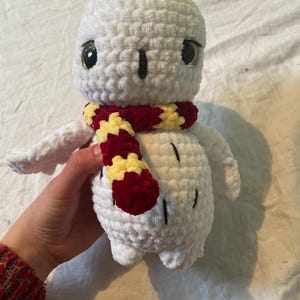 May include: A handmade white crochet owl plushie with a red and yellow scarf. The owl has large, golden eyes and black accents. The plushie is soft and cuddly, perfect for a child's toy or a decorative item.
