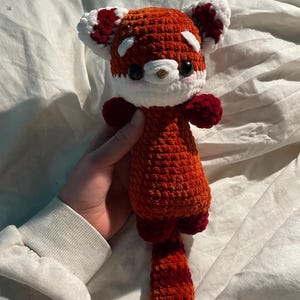 Op de afbeelding: Een handgemaakte gehaakte rode panda knuffel met een witte snuit, oren en accenten. Het knuffeldier heeft een lange staart en kleine ledematen. De rode panda wordt in een hand gehouden.