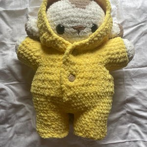 Può includere: Un orsacchiotto di peluche fatto a mano con una tuta con cappuccio gialla. L'orso ha una faccia bianca con macchie marroni, occhi neri e un piccolo naso marrone. La tuta è abbottonata con un bottone di legno.