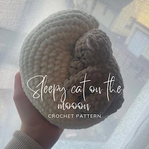 Könnte beinhalten: Eine gehäkelte Katze, die sich in einem größeren gehäkelten Halbmond befindet. Die Katze ist hellbraun und der Mond ist cremeweiß. Der Text "Sleepy cat on the moon" und "CROCHET PATTERN" ist auf dem Bild geschrieben.