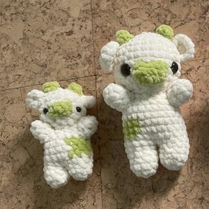 Könnte beinhalten: Zwei handgefertigte gehäkelte Kuh-Plüschtiere. Die Kühe sind weiß mit grünen Akzenten an Ohren, Nasen und Flecken. Sie haben schwarze Sicherheitsaugen und sind aus weichem, strukturiertem Garn gefertigt. Eines ist größer als das andere.