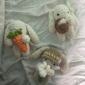 Könnte beinhalten: Drei handgefertigte Häkelhasen in Weiß, Beige und Braun. Ein Hase hält eine orangefarbene Karotte mit grünen Blättern, ein anderer einen braunen Korb und der dritte eine weiße Blume. Die Hasen haben lange Ohren.