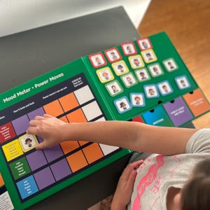 Puede incluir: Un juego de mesa verde y blanco "Mood Meter + Power Moves". El juego presenta una cuadrícula con etiquetas de emociones y una mano colocando una tarjeta en la emoción "Aterrado". El juego incluye tarjetas con diferentes expresiones faciales.