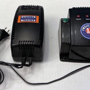 May include: Two black Lionel Trains power supply units. One unit has the Lionel Trains logo in an orange rectangle. The other unit has the Lionel logo in a blue circle, with an orange accent. A black power cord is connected to the units.