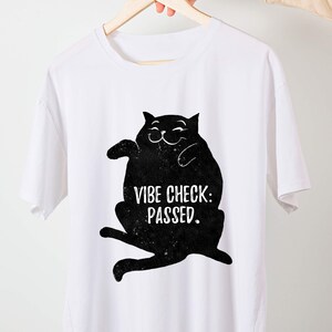 May include: White t-shirt featuring a black cat illustration with the text "VIBE CHECK: PASSED." The cat is smiling and has a relaxed pose. The shirt is made of a soft material and is suitable for casual wear.