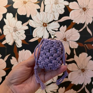 May include: A small, lavender macrame pouch with a silver-colored clasp. The pouch is held by a hand, and the background features a floral pattern with white and peach-colored flowers on a dark background.