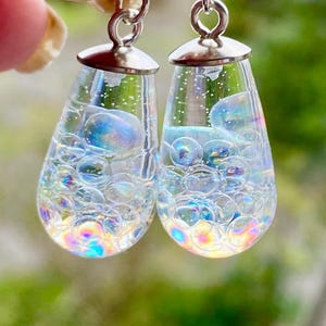 Könnte beinhalten: Ein Paar klare, tropfenförmige Ohrringe, gefüllt mit schillernden Blasen. Die Ohrringe haben silberfarbene Kappen und Haken. Die Blasen reflektieren einen Regenbogen von Farben und erzeugen einen schimmernden Effekt.