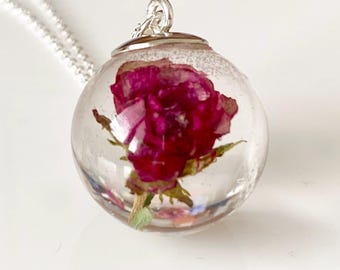 May include: A silver chain necklace with a clear, spherical pendant. The pendant contains a preserved, deep red rose. The necklace is set against a white background, with small pink and blue flowers scattered around.