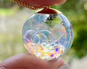 May include: A clear, spherical pendant necklace with a gold chain. The pendant contains iridescent, bubble-like shapes. The gold chain is a delicate, linked design. The pendant is held up by a person's fingers.