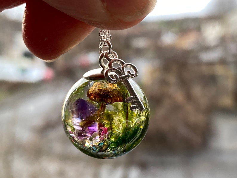 May include: A silver necklace with a clear glass orb pendant containing a miniature mushroom, purple flowers, and green moss. A silver key charm hangs from the orb.