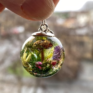 May include: A silver pendant featuring a clear glass sphere filled with a miniature forest scene, including a small mushroom, green moss, and purple and pink flowers.