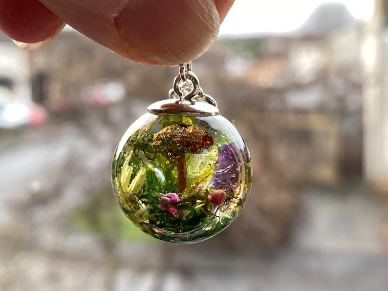 May include: A silver pendant featuring a clear glass sphere filled with a miniature forest scene, including a small mushroom, green moss, and purple and pink flowers.