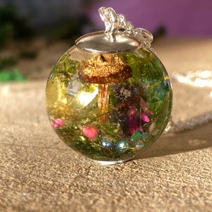 May include: A silver chain necklace with a round glass pendant containing a miniature forest scene. The pendant features a small mushroom, green moss, and pink flowers.