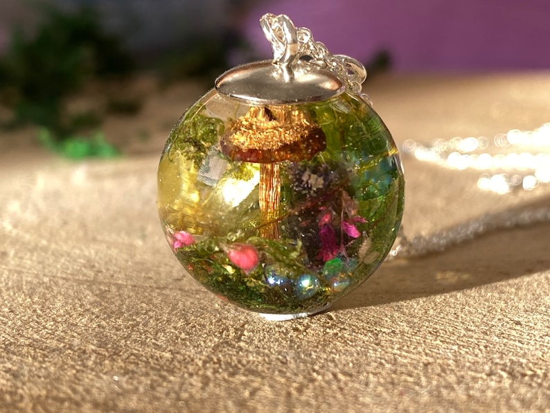 May include: A silver chain necklace with a round glass pendant containing a miniature forest scene. The pendant features a small mushroom, green moss, and pink flowers.