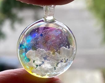 May include: A clear, round pendant with a glass loop. The pendant contains a colorful, swirling design with blue, purple, and yellow hues, and white cloud-like shapes. The pendant is held between two fingers.