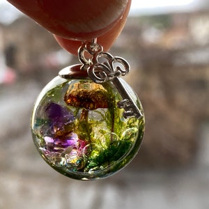 May include: A clear glass sphere pendant with a silver key charm. Inside the sphere are preserved green moss, purple flowers, and a brown mushroom.