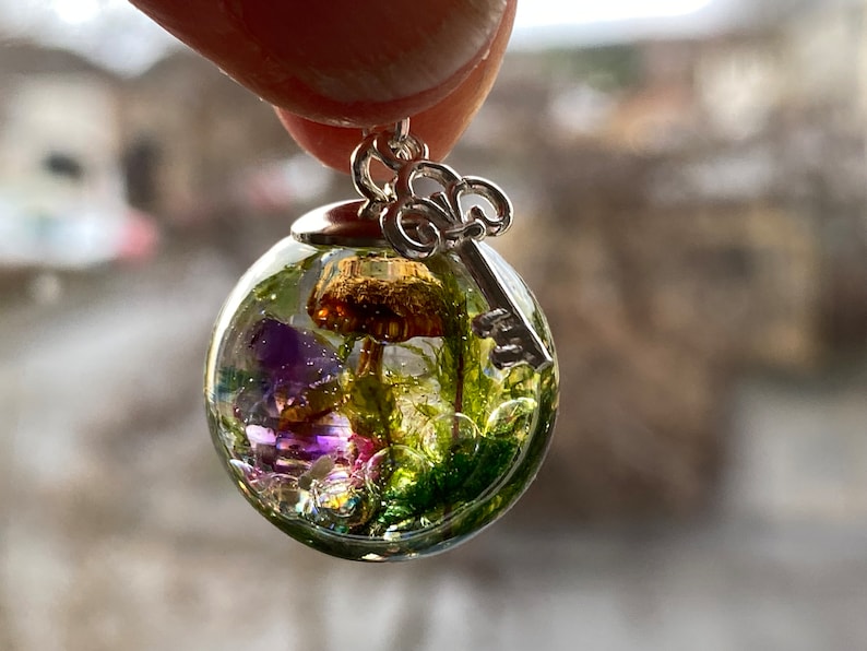 May include: A clear glass sphere pendant with a silver key charm. Inside the sphere are preserved green moss, purple flowers, and a brown mushroom.
