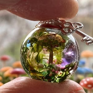 May include: A clear glass sphere pendant necklace with a silver key charm. Inside the sphere is a miniature forest scene with a small tree, green moss, and purple flowers.