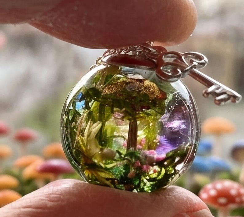 May include: A clear glass sphere pendant necklace with a silver key charm. Inside the sphere is a miniature forest scene with a small tree, green moss, and purple flowers.