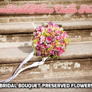 Puede incluir: Un ramo de novia de flores preservadas en tonos rosas y verdes, con una cinta blanca, sobre un conjunto de escalones de piedra. El texto "Your Bridal Flowers Custom Necklace" se muestra sobre el ramo.