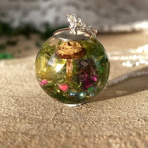 May include: A silver chain necklace with a round glass pendant containing a miniature forest scene. The pendant features a small mushroom, green moss, and pink flowers.