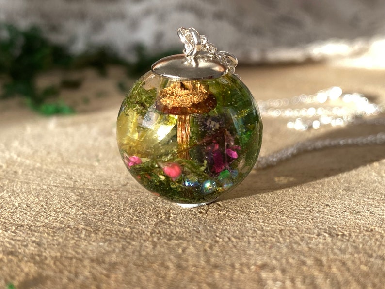 May include: A silver chain necklace with a round glass pendant containing a miniature forest scene. The pendant features a small mushroom, green moss, and pink flowers.