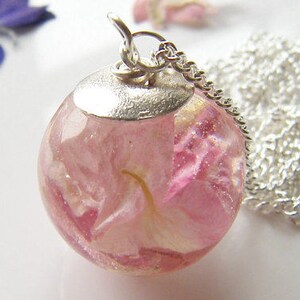 Puede incluir: Un collar de cadena de plata con un colgante redondo que contiene una flor rosa prensada en su interior. El colgante es transparente y la flor es visible.