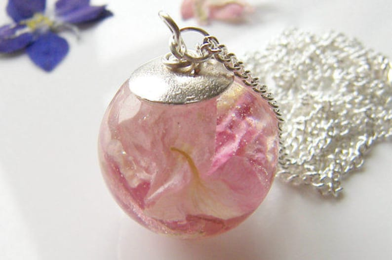Puede incluir: Un collar de cadena de plata con un colgante redondo que contiene una flor rosa prensada en su interior. El colgante es transparente y la flor es visible.