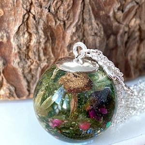 May include: A silver chain necklace with a round glass pendant containing a preserved mushroom and other botanical elements. The pendant is filled with green moss, pink flowers, and glitter.