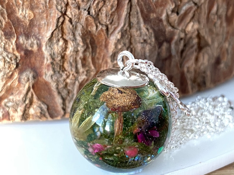 May include: A silver chain necklace with a round glass pendant containing a preserved mushroom and other botanical elements. The pendant is filled with green moss, pink flowers, and glitter.