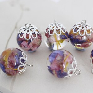 Puede incluir: Seis colgantes de plata con delicados dise&ntilde;os florales. Cada colgante presenta una esfera de vidrio transparente llena de flores prensadas en tonos de rosa, morado y amarillo.