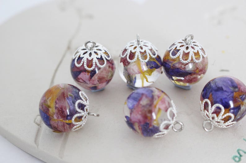 Puede incluir: Seis colgantes de plata con delicados dise&ntilde;os florales. Cada colgante presenta una esfera de vidrio transparente llena de flores prensadas en tonos de rosa, morado y amarillo.