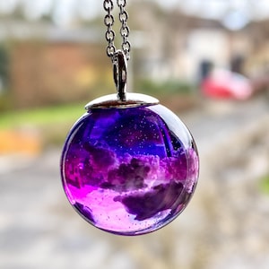 Può includere: Una collana con ciondolo sferico caratterizzata da un globo in resina trasparente riempito con sfumature vorticose di viola intenso e magenta vibrante, che ricorda una nebulosa. Il globo è sormontato da una montatura in metallo argentato.