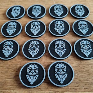 Könnte beinhalten: Eine Ansammlung von zwölf schwarzen und grauen runden Token. Jeder Token zeigt ein stilisiertes, bärtiges Gesichtsdesign in Grau, vor einem schwarzen Hintergrund mit einem grauen Rand. Die Token sind auf einer Holzoberfläche angeordnet.