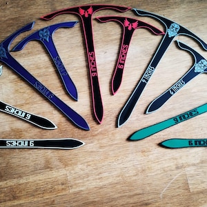 Op de afbeelding: Een verzameling decoratieve T-vormige gereedschappen in zwart, blauw, rood en groen, elk met een sierlijk ontwerp. De gereedschappen zijn gemarkeerd met "6 INCHES" (15,24 cm) of "9 INCHES" (22,86 cm) en liggen op een houten oppervlak.