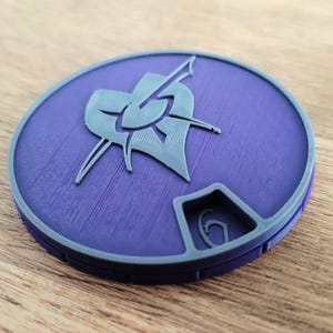 Peut inclure: Un emblème rond, violet et gris, avec un motif stylisé. Il présente un motif central en forme de bouclier et d'épée. Le bord extérieur est gris, et le fond est violet. L'objet est imprimé en 3D.