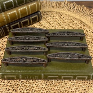 以下が含まれることがあります： 緑色の本の上に配置された、花と葉のディテールが施された7つのブロンズ色のキャビネットプル。 プルは湾曲した形状で、短いポストに取り付けられています。 背景には、本の山と織りのマットが含まれています。