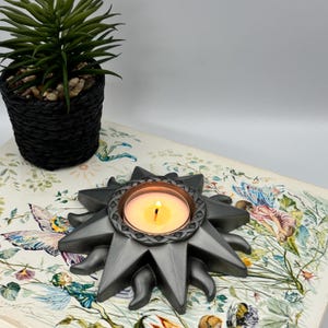 Może przedstawiać: Ciemnoszary świecznik w kształcie słońca z centralnym okrągłym otworem, w którym znajduje się zapalona świeczka typu tealight. Świecznik posiada teksturowaną, plecioną obwódkę wokół środka oraz promienie w kształcie gwiazdy.