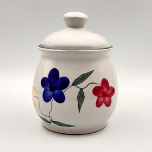Peut inclure: Un pot en céramique blanc avec un couvercle, décoré de fleurs peintes à la main en bleu, rouge et jaune. Le pot a une forme arrondie et un petit bouton sur le couvercle. Le motif floral est complété par des feuilles vertes.