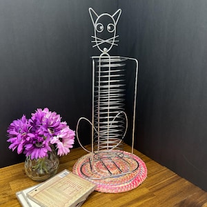 May include: A silver-colored wire cat-shaped CD rack with a round base. The rack holds CDs in a vertical position. The base is a woven pink and red mat. A vase of purple flowers sits to the left of the rack.