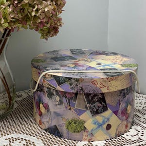 May include: A round decorative box with a lid, featuring a collage of vintage images, letters, and floral elements in shades of purple, cream, and yellow. The box has a white rope handle and is placed on a white lace tablecloth.