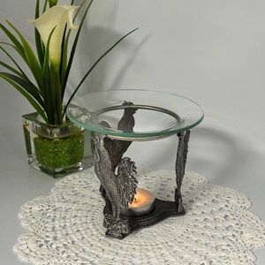 May include: A metal oil warmer featuring three sculpted rooster figures supporting a circular clear glass dish. The base is triangular, and a small tea light candle is positioned in the centre, emitting a warm glow beneath the glass bowl.